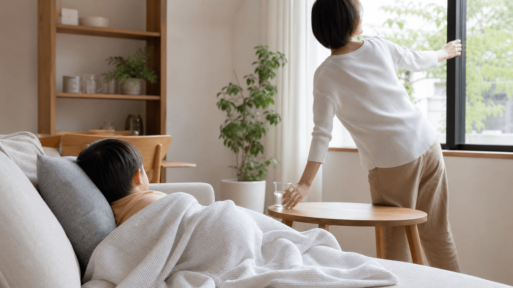 リビングのソファで休む子どものそばで保護者が窓を開けている場面