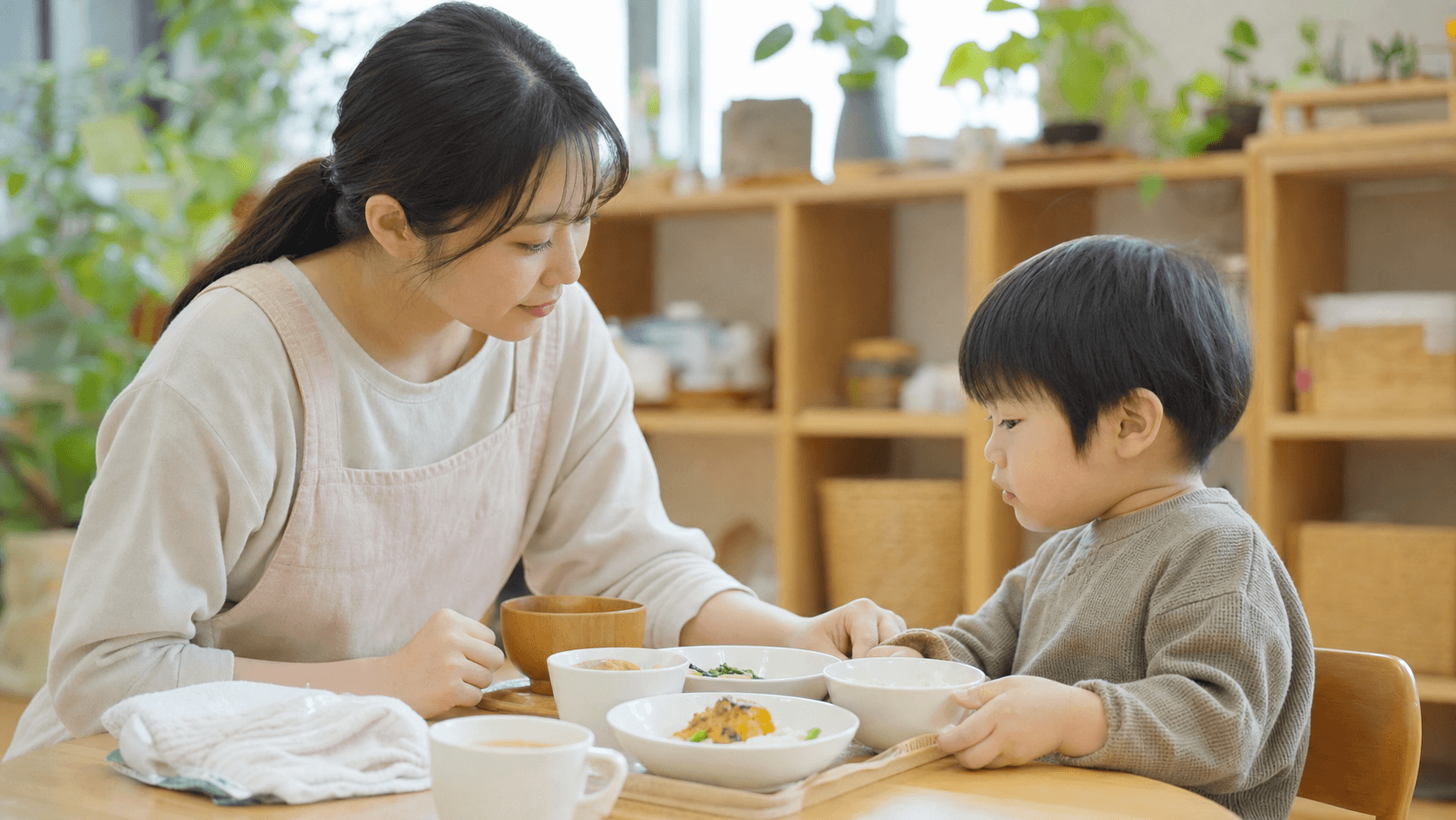 保育室の食卓で保育者が子どもの食事を一緒に確認している場面