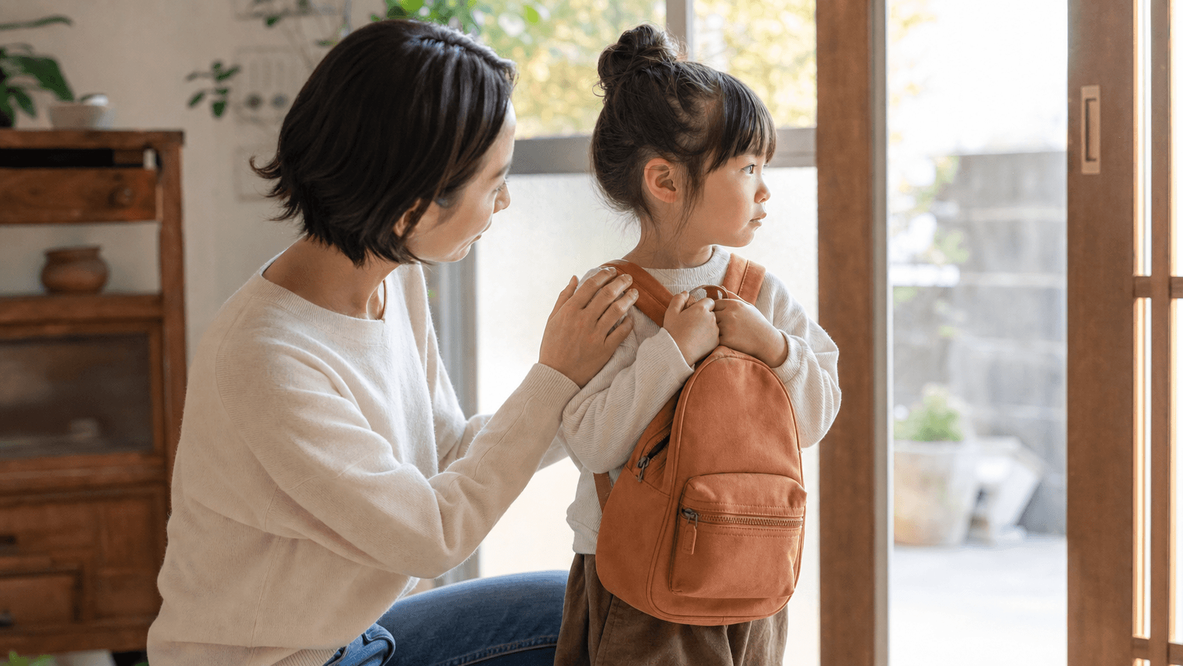 玄関で通園バッグを持つ子どもに保護者が寄り添っている場面