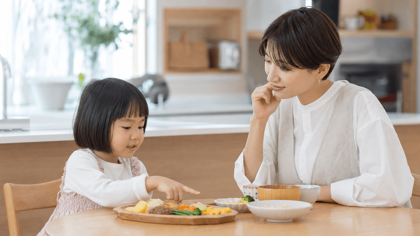 食卓で子どもが食べたいものを指し保護者が見守っている場面